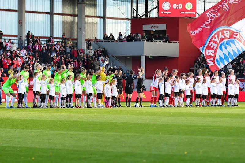 Die Mannschaften vom FC Bayern München und VfL Wolfsburg vor einem Spiel der Frauen-Bundesliga. Die Teams stehen mit den Einlaufkindern in einer Reihe und winken in Richtung des Publikums.