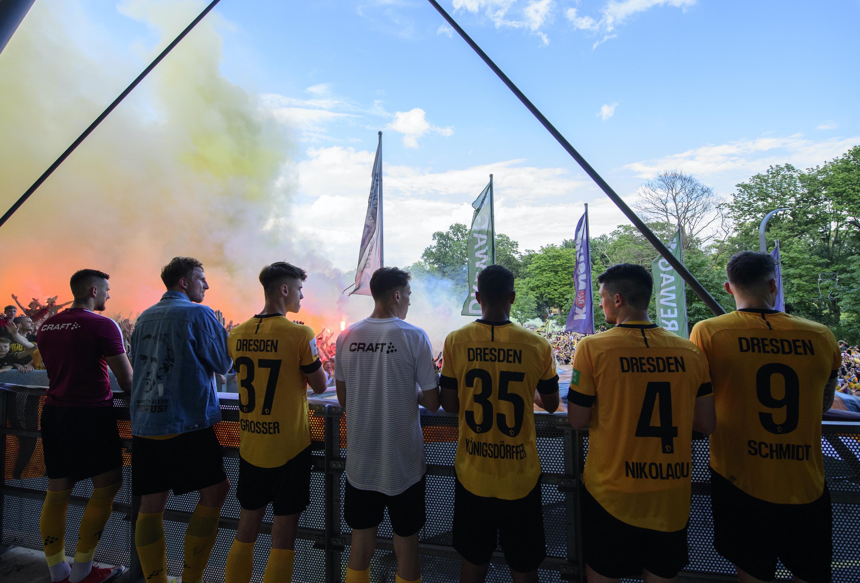 Die Mannschaft von Dynamo Dresden bei Feierlichkeiten nach dem Aufstieg. Im Hintergrund sind Fans und gelber Rauch zu sehen.